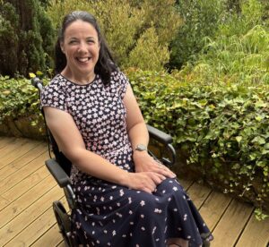 Aideen sat smiling, sitting in her manual wheelchair with trees and greenery behind her