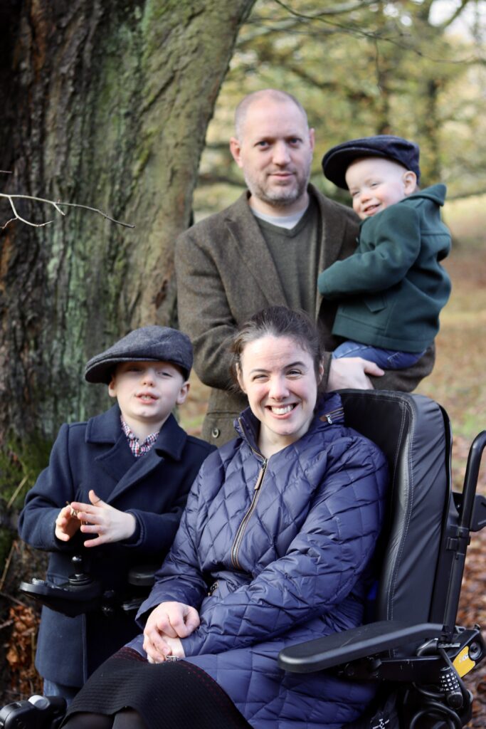 Aideen Blackborough with her husband and two sons outdoors in autumn woodland. Aideen is an adult living with cerebral palsy.